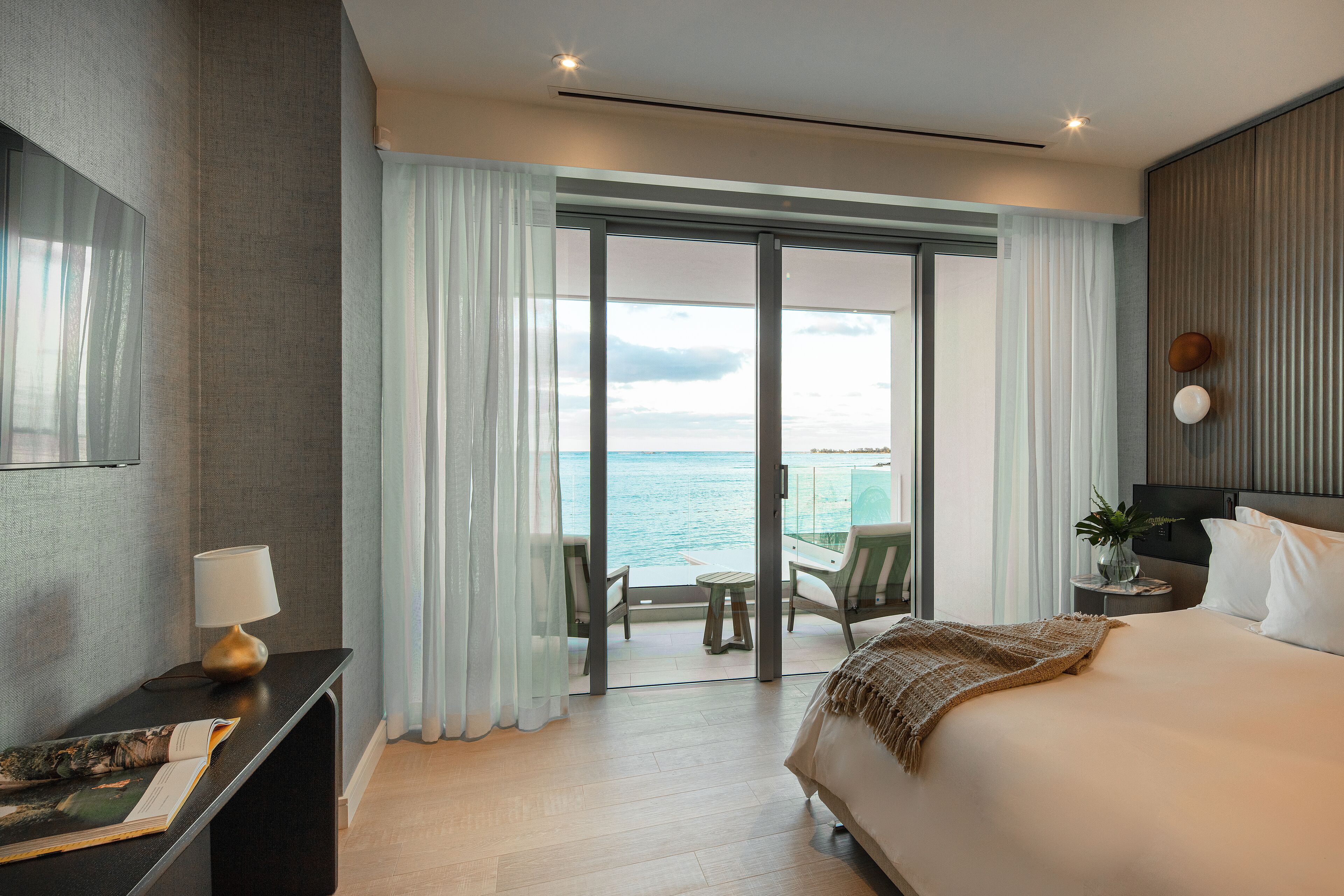 A hotel room with a large bed, a desk with a lamp, a view of the sea through a sliding door, and a small table with chairs outside.