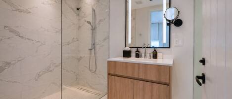 Un baño moderno con ducha acristalada, tocador de madera con espejo y suelo de baldosas.