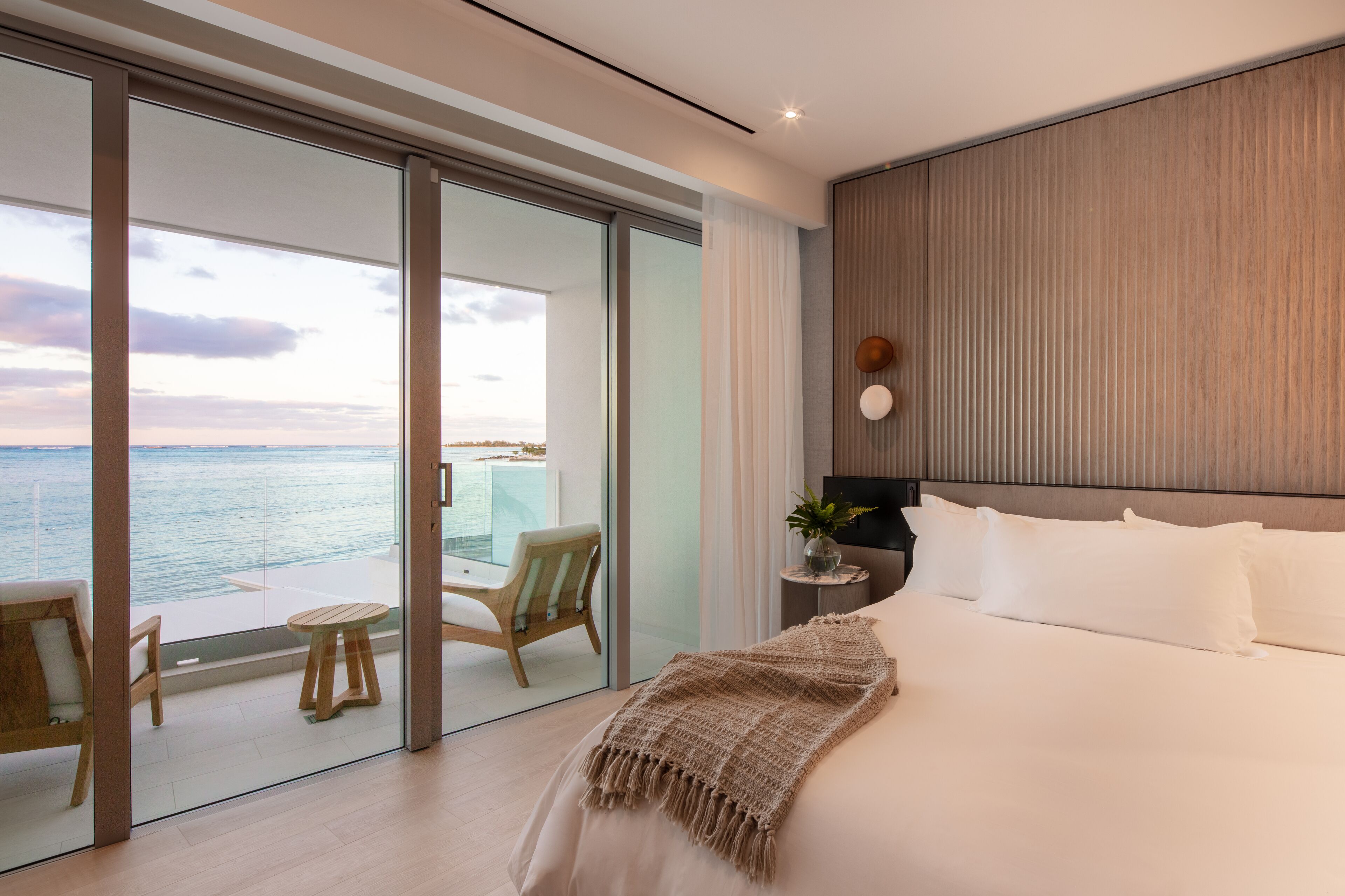 Una habitación de hotel moderna con una cama grande, vistas al mar a través de puertas de vidrio del piso al techo, un balcón pequeño con una mesa y sillas, y una mesita de noche con una planta.