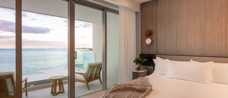 Una habitación de hotel moderna con una cama grande, vistas al mar a través de puertas de vidrio del piso al techo, un balcón pequeño con una mesa y sillas, y una mesita de noche con una planta.