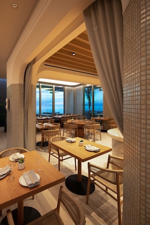 A modern dining area with wooden tables and chairs, large windows offering a view of the sea, and a tiled wall on one side.