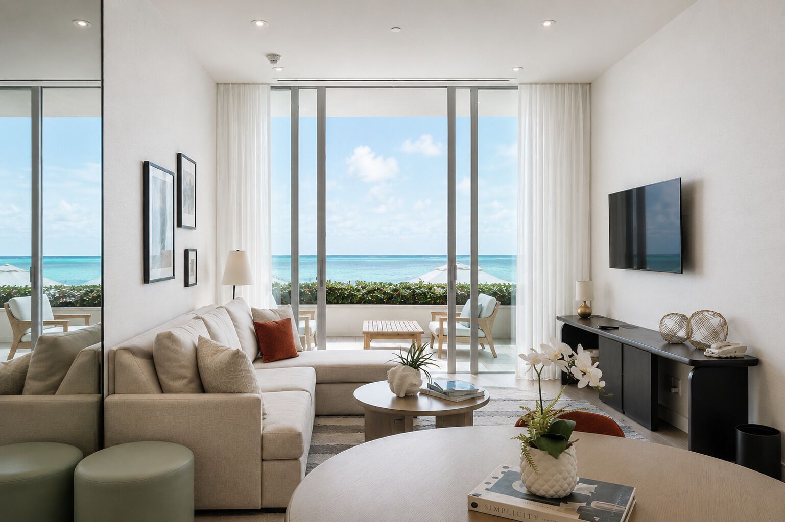 A modern living room with a large window overlooking the ocean, a flat-screen TV mounted on the wall, and a cozy seating area with a sofa and armchairs.