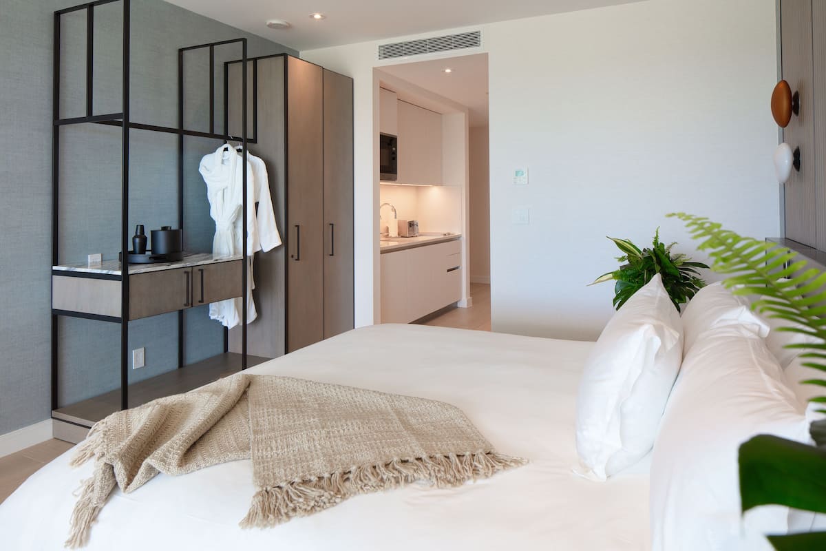 A modern bedroom with a large bed, a bedside table, a wardrobe, and a kitchenette in the background.