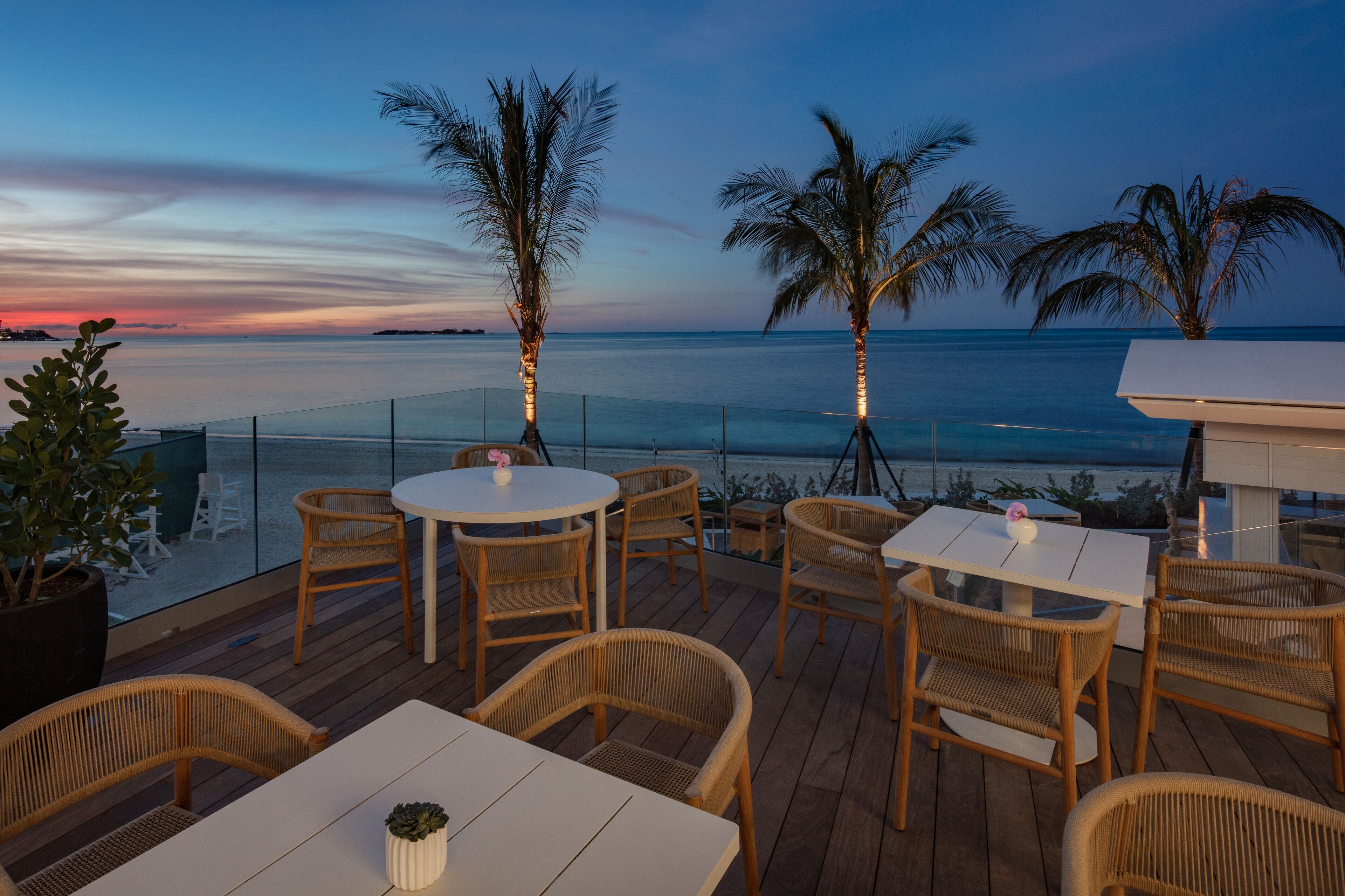 A wooden deck with round tables and wicker chairs, overlooking a calm sea at dusk.