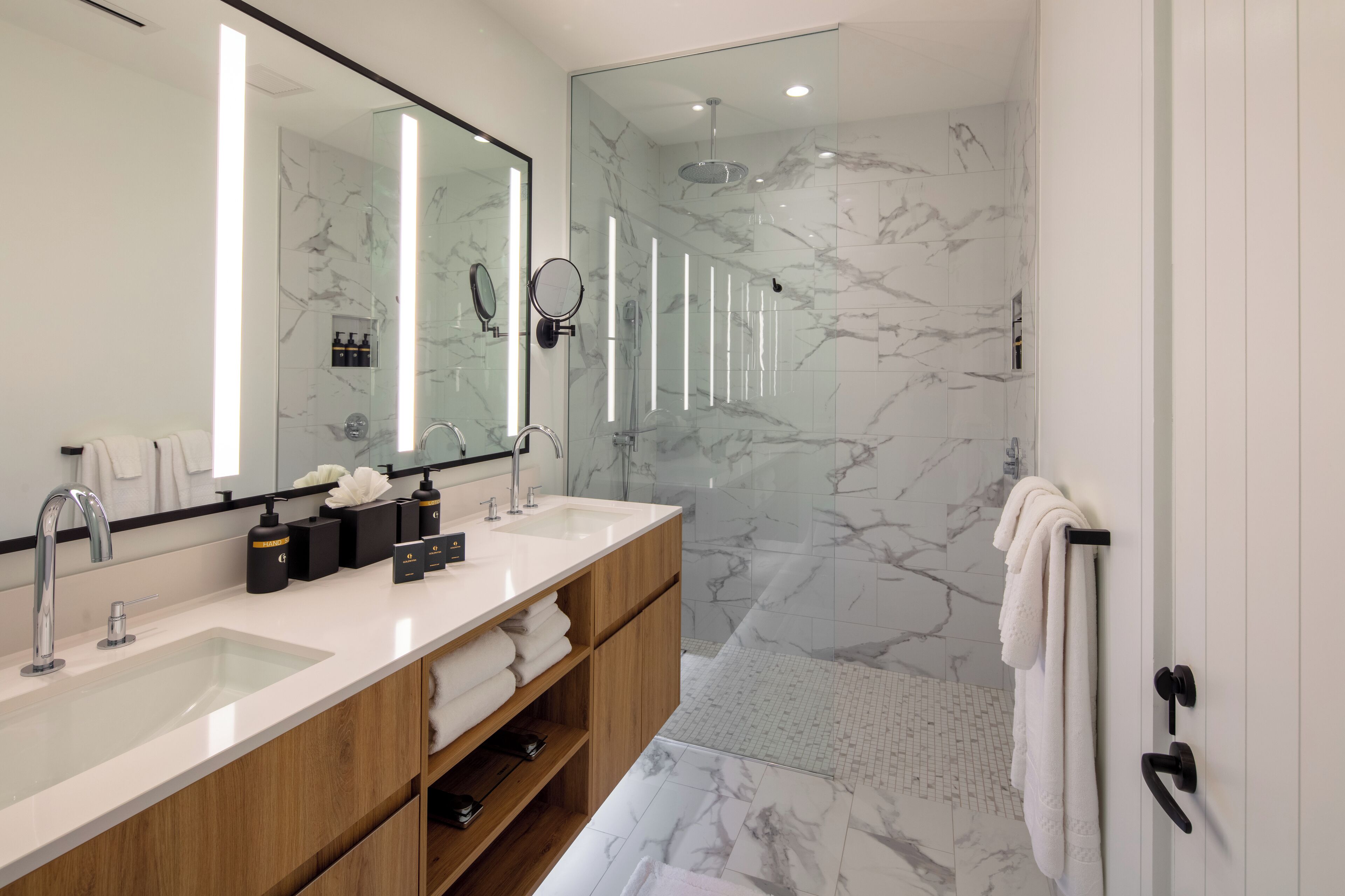 A modern bathroom with a large mirror, two sinks, a shower, and marble flooring.
