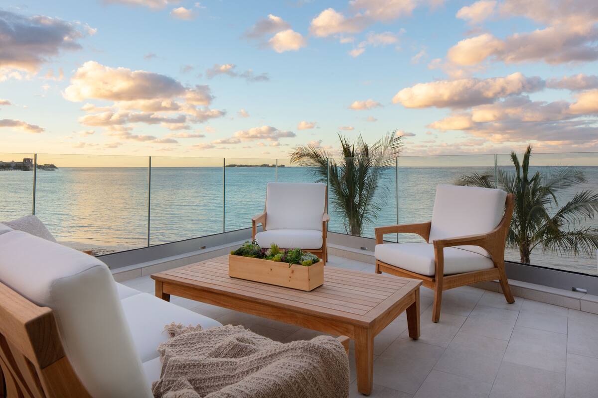A balcony with a view of the sea, featuring a wooden table, two white cushioned chairs, and a small plant arrangement.