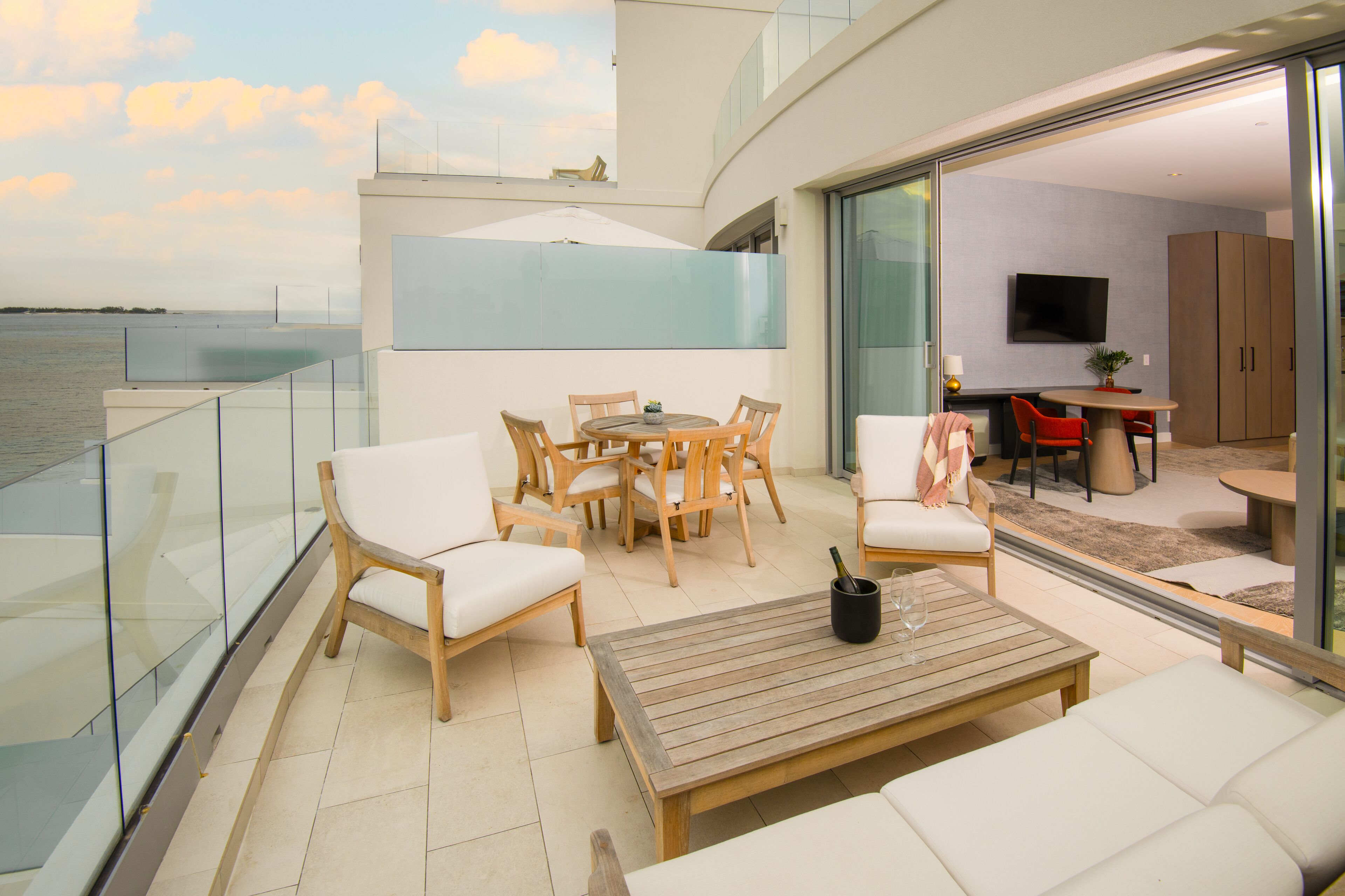 A modern outdoor living area with a wooden table, white cushioned chairs, and a glass balustrade overlooking a body of water.