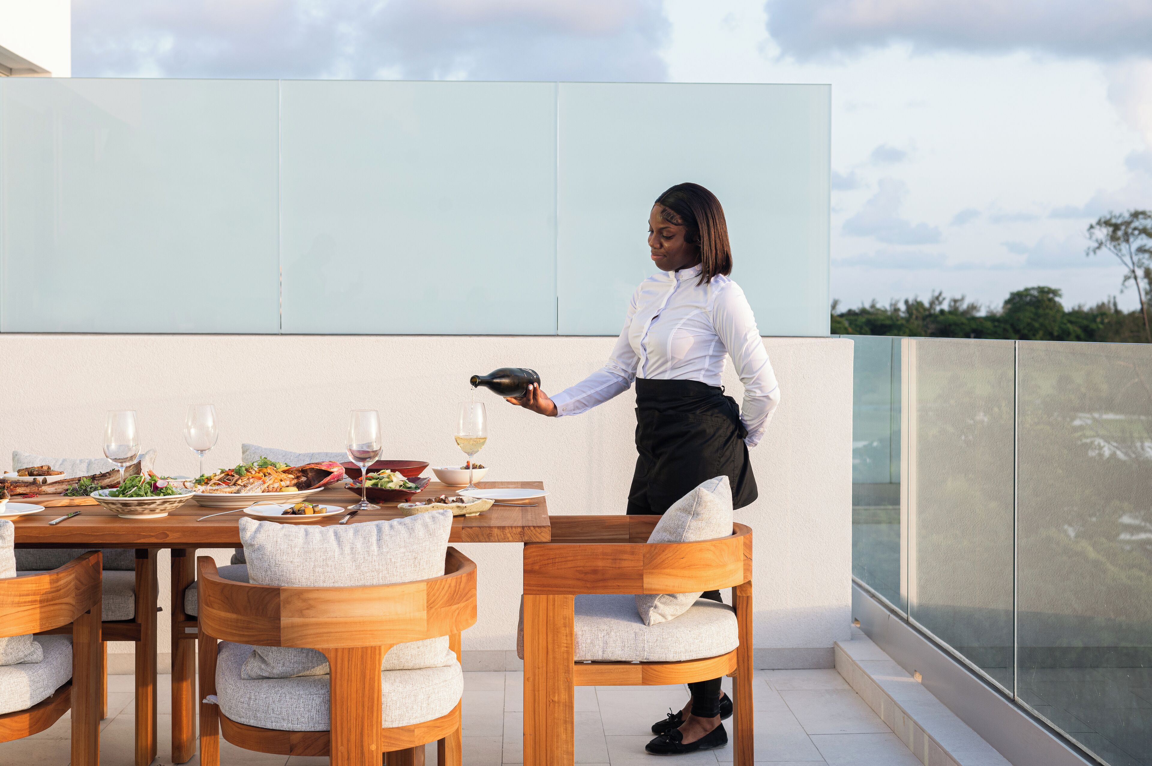 Un comedor con una mesa dispuesta para dos personas, un camarero sirviendo y un edificio moderno al fondo.