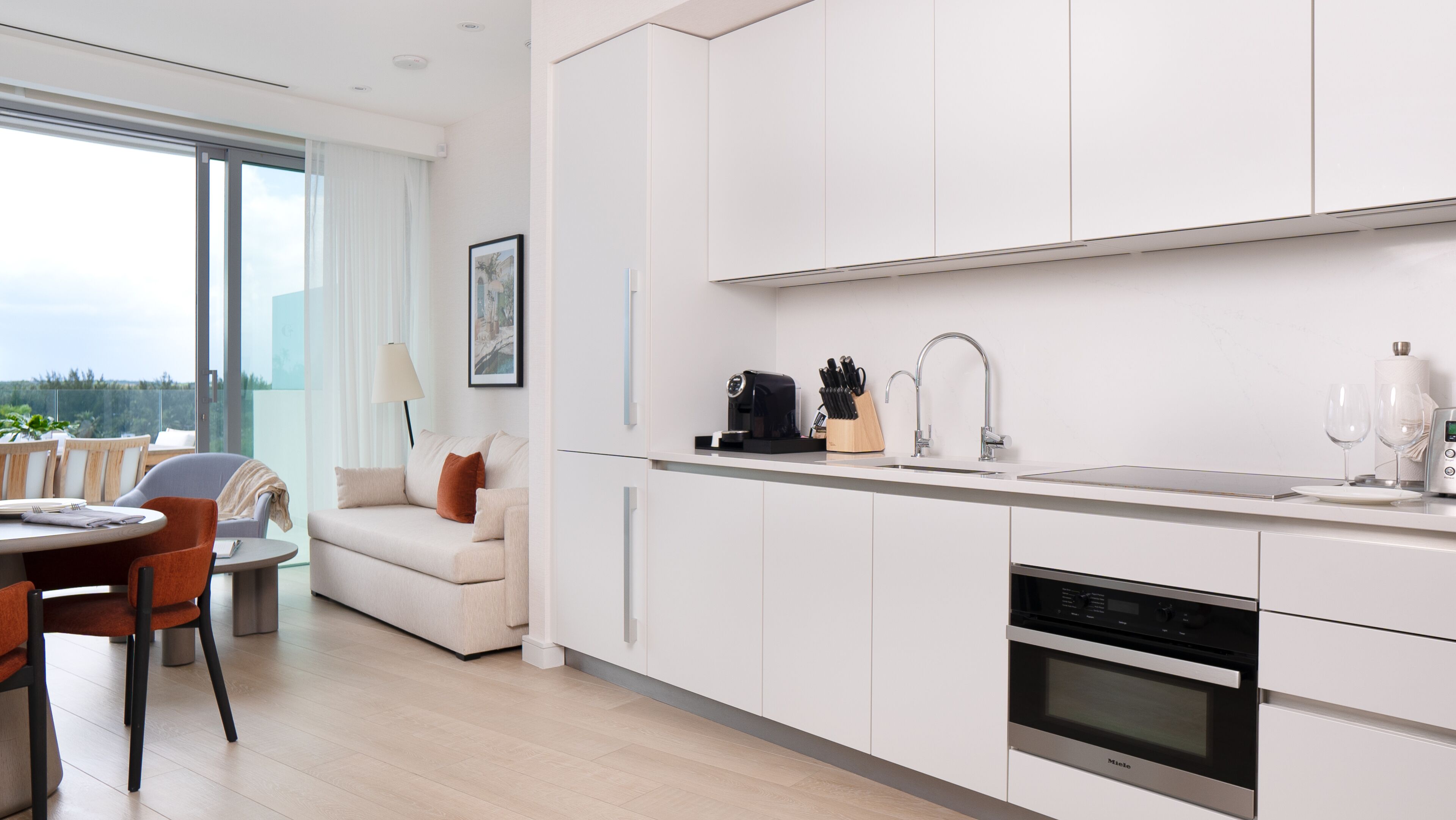 A modern kitchen with white cabinets, a coffee machine, and a sink. There is a dining area with a table and chairs, and a sofa.
