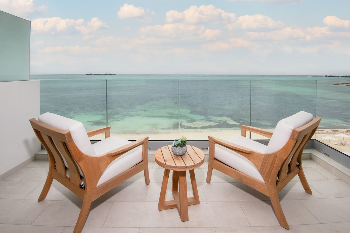 A balcony with two wooden chairs and a small table, overlooking a clear blue sea and a distant island.