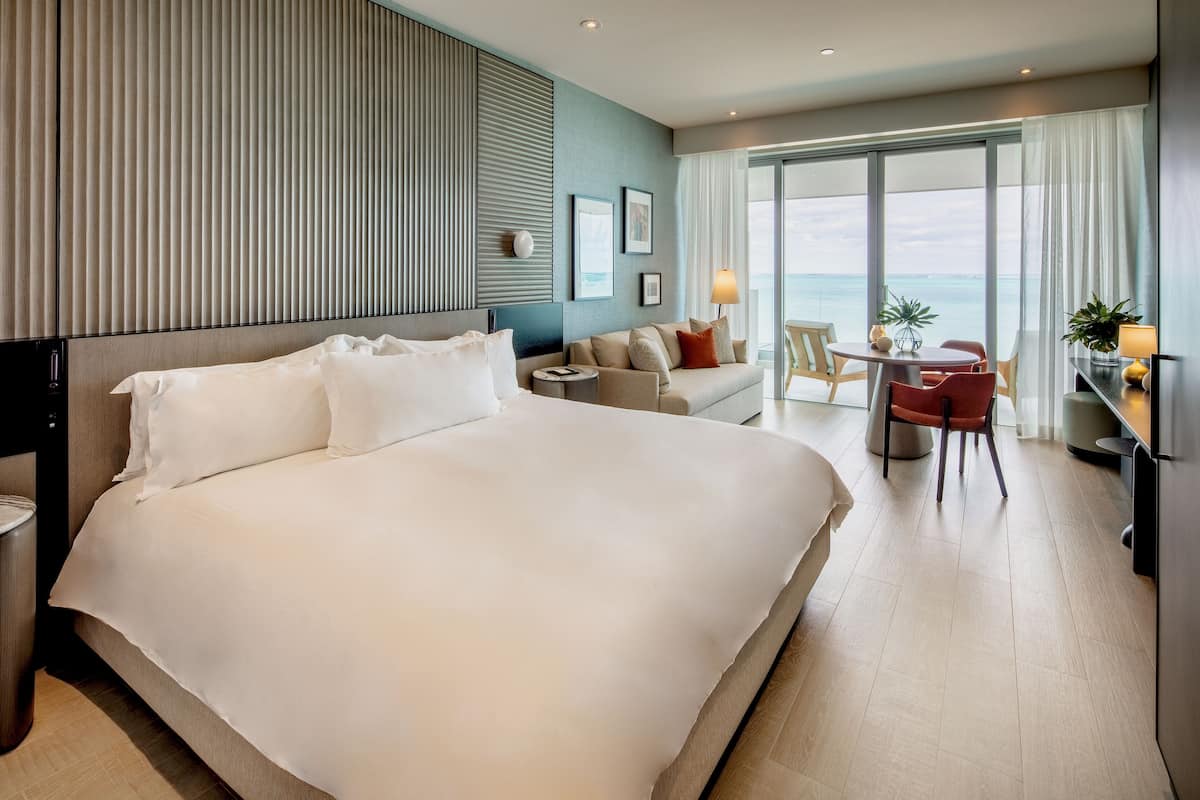 A hotel room with a large bed, a sofa, a small table with chairs, a lamp, and a view of the sea through large windows.