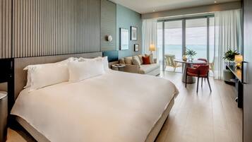 A hotel room with a large bed, a sofa, a small table with chairs, a lamp, and a view of the sea through large windows.