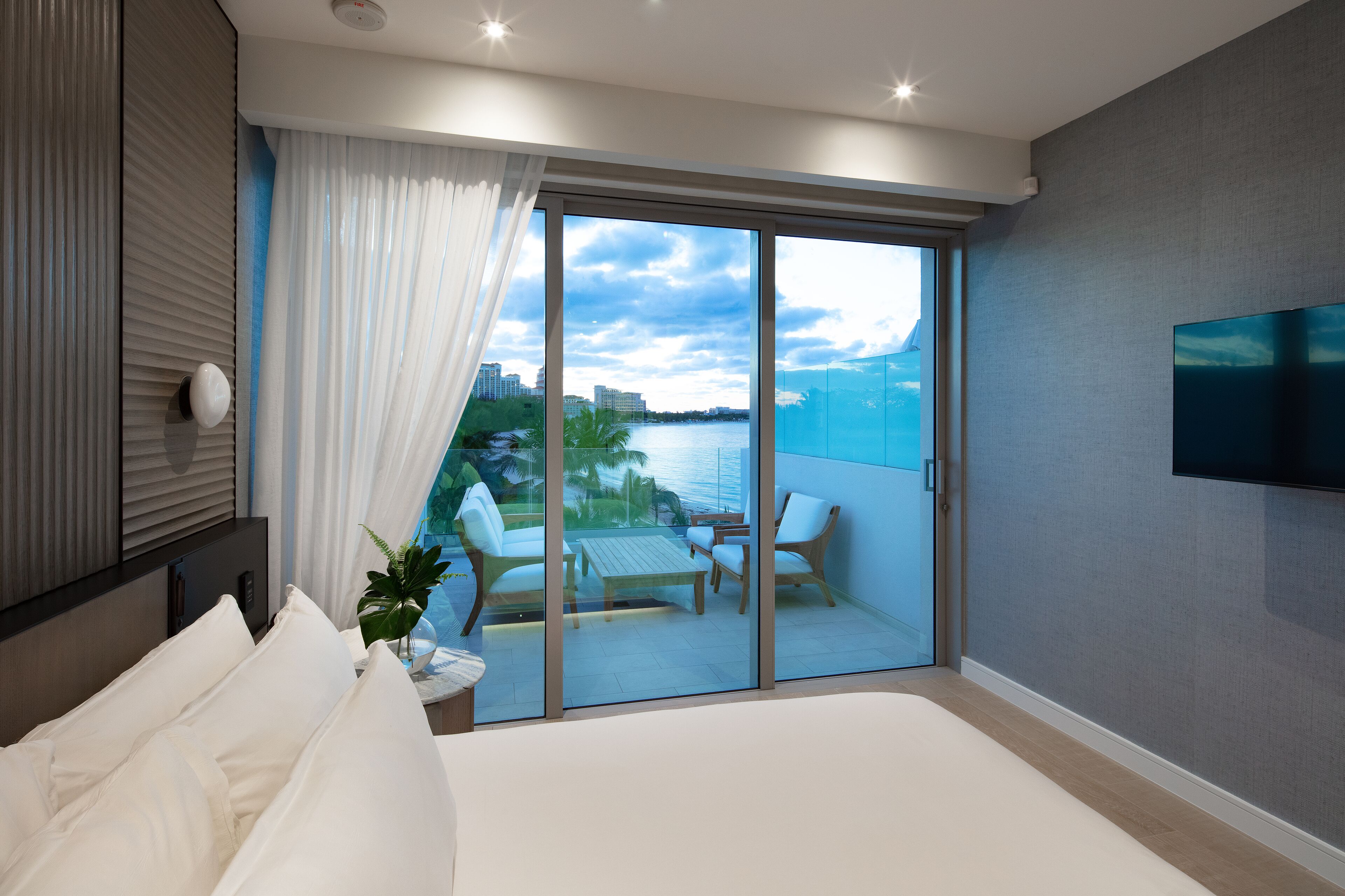 Una habitación de hotel moderna con una cama grande, un balcón con mesa y sillas, un televisor montado en la pared y vistas al mar y edificios.