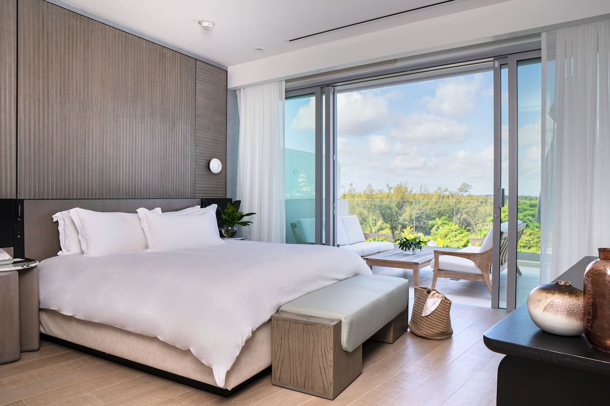 A modern hotel room with a large bed, a small table, a bench, and a view of the outdoors.