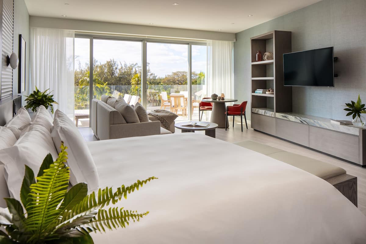 A modern hotel room with a large bed, a TV mounted on the wall, a balcony with a dining area, and a bookshelf.