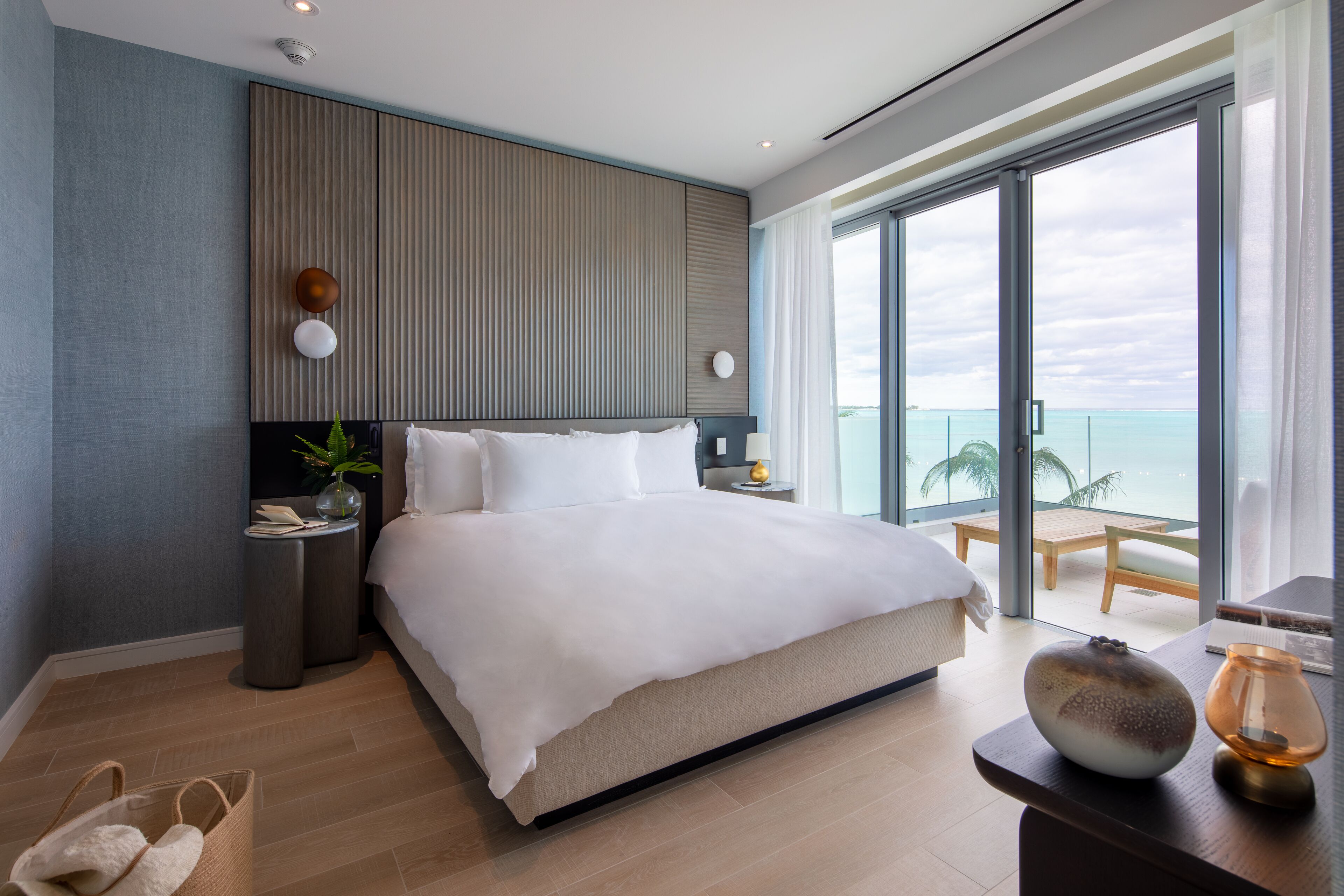 A modern hotel room with a large bed, a nightstand, a bedside table with a lamp, a view of the ocean, and a balcony with a table and chairs.