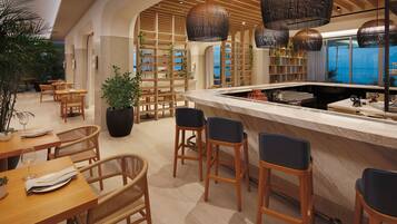 A modern restaurant with wooden furniture, a central bar area, and large windows offering a view of the sea.