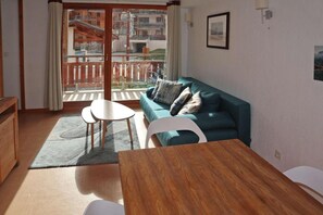 En stue med sofa, to små borde, et træsofabord og udsigt til en altan med gelænder samt en bygning i baggrunden.