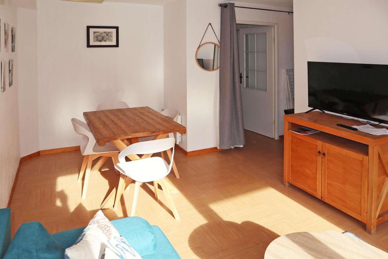 A living room with a wooden dining table, white chairs, a TV on a wooden stand, and a door with a grey curtain.