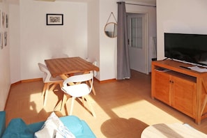 A living room with a wooden dining table, white chairs, a TV on a wooden stand, and a door with a grey curtain. - Ski-in/ski-out & foot of the slopes, 1st floor, swimming pool, sauna, balcony, tv, ski locker, 34m² (Les Orres)