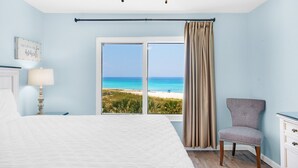 A bedroom with a bed, a chair, a nightstand, a lamp, a window with a beach view, and a ceiling fan. - ✨FAB Gulf Views~Pool~Beach Svc~2BR Destin on the Gulf 402 (Destin)