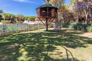 Una casa del árbol de madera con techo verde, construida sobre un único tronco, rodeada por un patio cercado con césped y árboles.