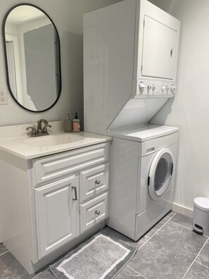 A modern bathroom with a built-in washing machine, a vanity with drawers, a mirror, and a small trash bin.