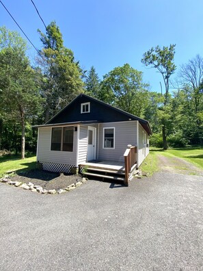 A small, single-story cabin with a front porch and a covered entryway, surrounded by a gravel driveway and a grassy yard.