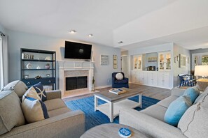 A modern living room with a fireplace, a television, and a kitchen area in the background. - Lagoon-Front Retreat w/ Pool, Spa, Game Room & Private Dock Short Walk to Beach (Hilton Head Island)