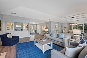 A spacious living room with a blue rug, a coffee table, and a grey sofa. There is a kitchen area with white cabinets and a dining area with a table and chairs. - Lagoon-Front Retreat w/ Pool, Spa, Game Room & Private Dock Short Walk to Beach (Hilton Head Island)