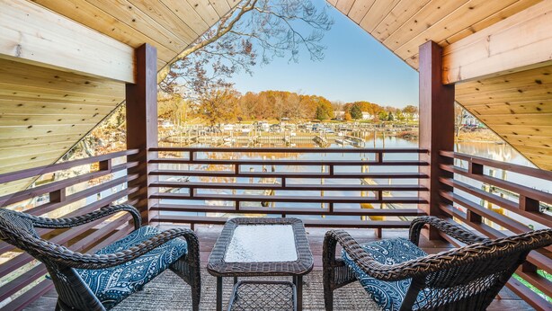 Une terrasse en bois, agrémentée de meubles en osier et d’une table, qui offre une vue sur une étendue d’eau et les arbres aux couleurs automnales.