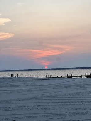 A beach with a wooden pier extending into calm waters, a sunset in the background, and a clear sky.