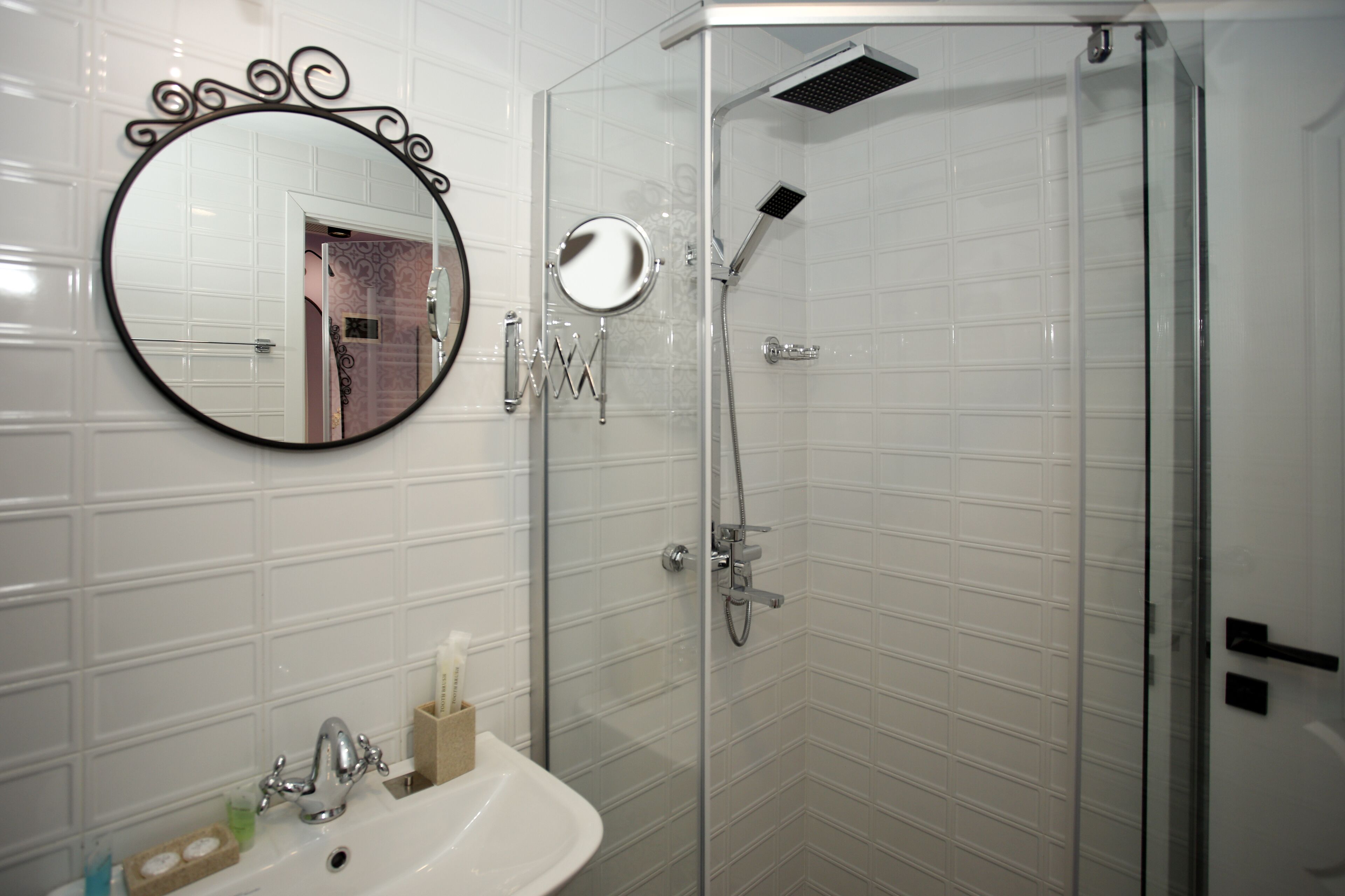 A bathroom with a white tiled wall, a round mirror with a decorative frame, a sink with a soap dish, and a glass-enclosed shower area.