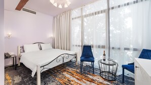 A hotel room with a bed, two chairs, a small table with a bottle and glass, and large windows with sheer curtains.
