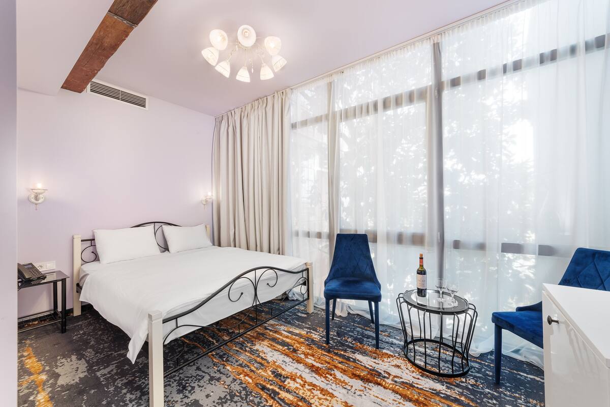 A hotel room with a bed, two chairs, a small table with a bottle and glass, and large windows with sheer curtains.