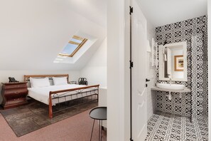 A bedroom with a skylight, a bed, a dresser, and a chair. The bathroom has patterned tiles and a sink with a mirror.