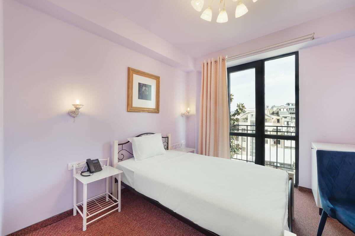 A hotel room with a bed, bedside table, lamp, and a view of buildings through a window.