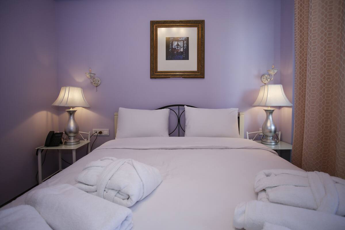 A hotel room with a bed, two bedside lamps, a framed picture on the wall, and two rolled towels on the bed.