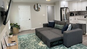 A modern living room with a grey sectional sofa, a flat-screen TV mounted on the wall, a ceiling fan, and a kitchen area with white cabinets and stainless steel appliances.