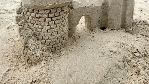 Un castell de sorra amb base de maó i part superior cònica, situat a una platja sorrenca amb l'oceà al fons.