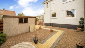 A residential property with a paved patio, a small garden area with potted plants, and a brick pathway leading to a door. - Beautiful Three Bedroom Seaside Retreat (Prestonpans)