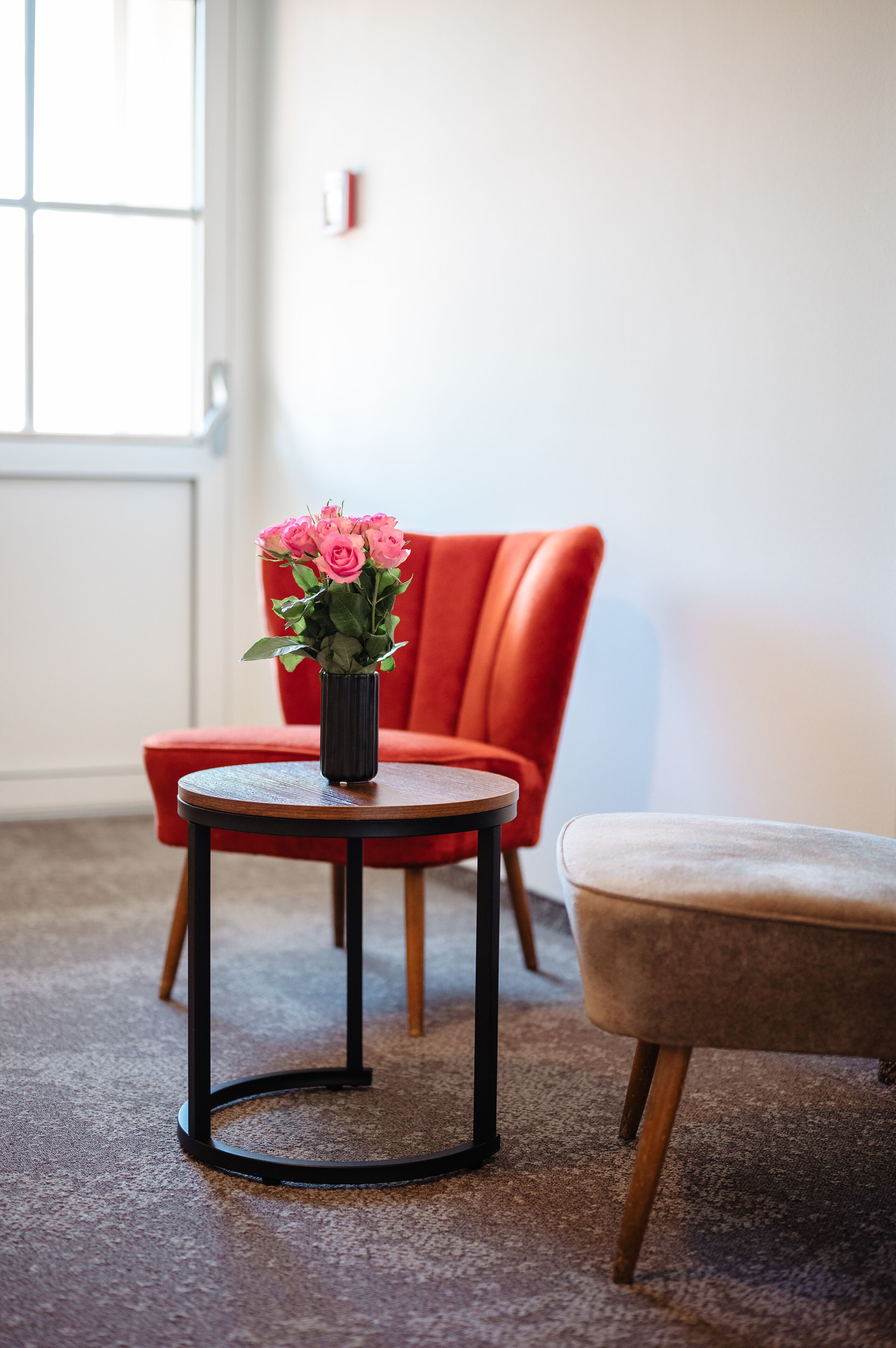 A room with a red chair, a small round table with a black vase of pink flowers, and a beige ottoman.