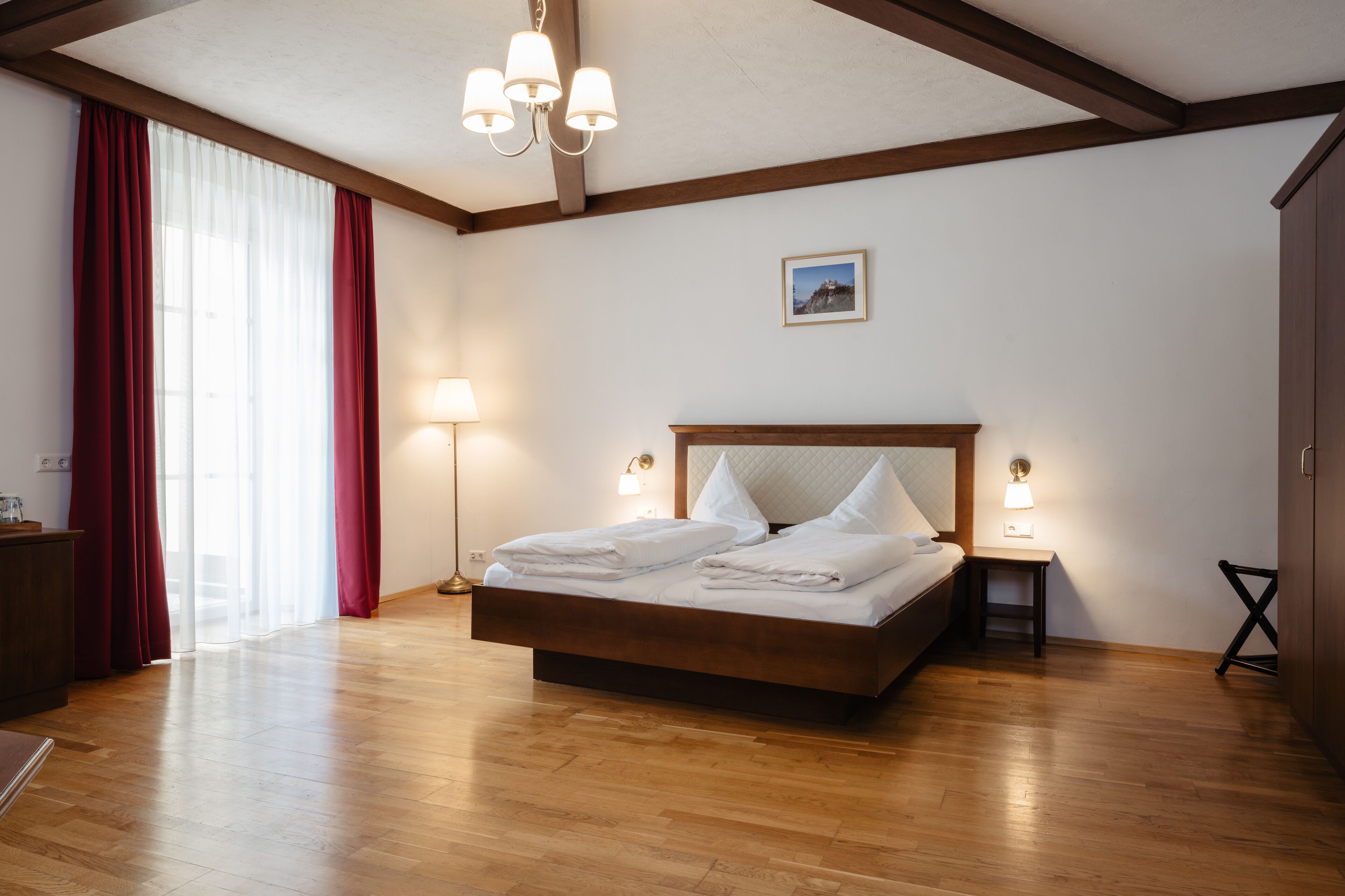 Un dormitorio con una cama de madera, una mesita de noche con lámpara, un escritorio con lámpara y un ventanal con cortinas rojas.