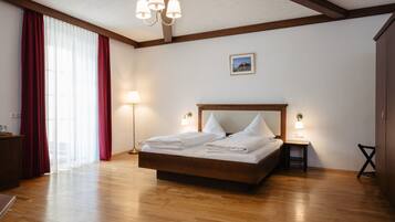 Un dormitorio con una cama de madera, una mesita de noche con lámpara, un escritorio con lámpara y un ventanal con cortinas rojas.