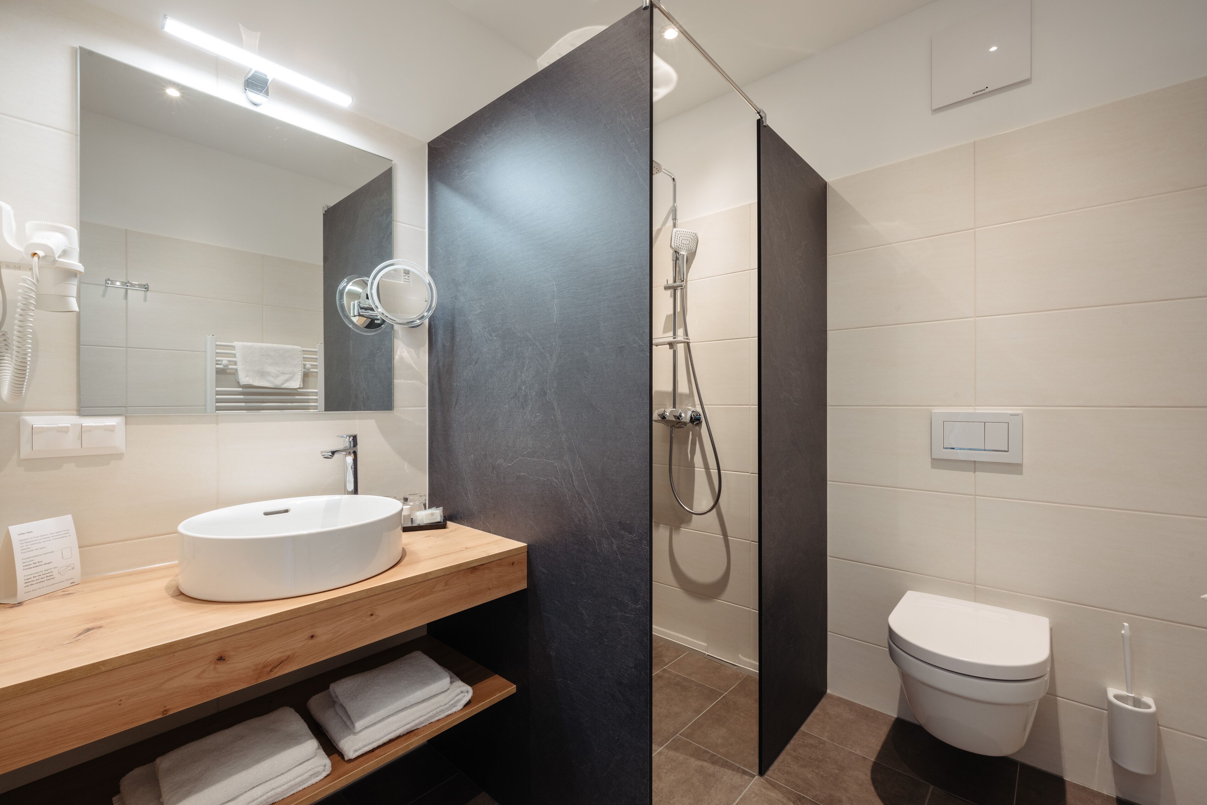 A modern bathroom with a wooden vanity, a white sink, a large mirror, a shower enclosure, and a toilet.