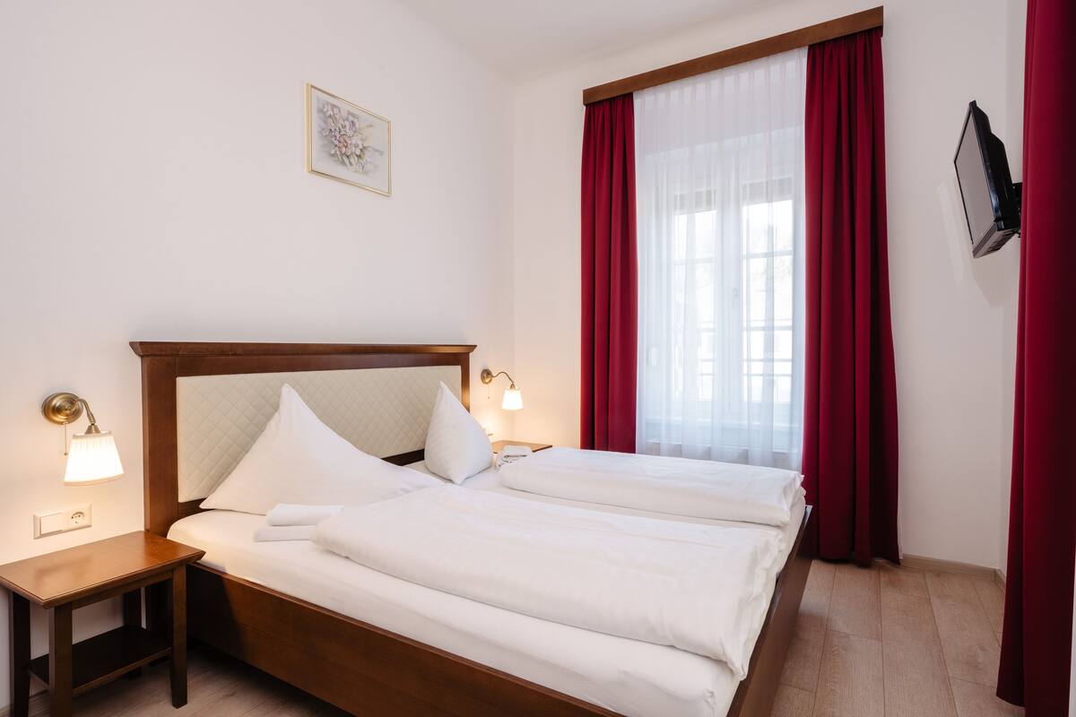Una habitación de hotel con una cama grande, dos mesitas de noche con lámparas, un televisor montado en la pared y una ventana con cortinas rojas.