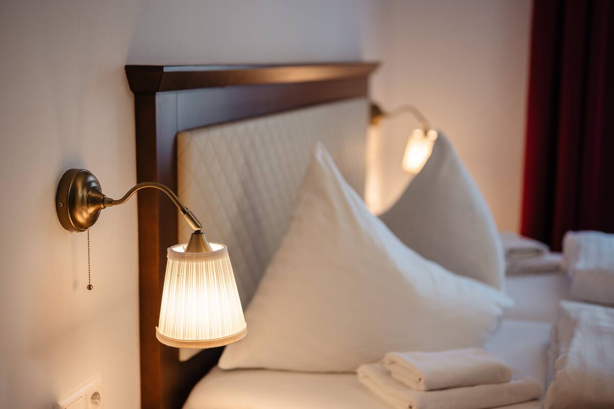 Una cama bien hecha con sábanas blancas, una lámpara de pared y un cabecero.