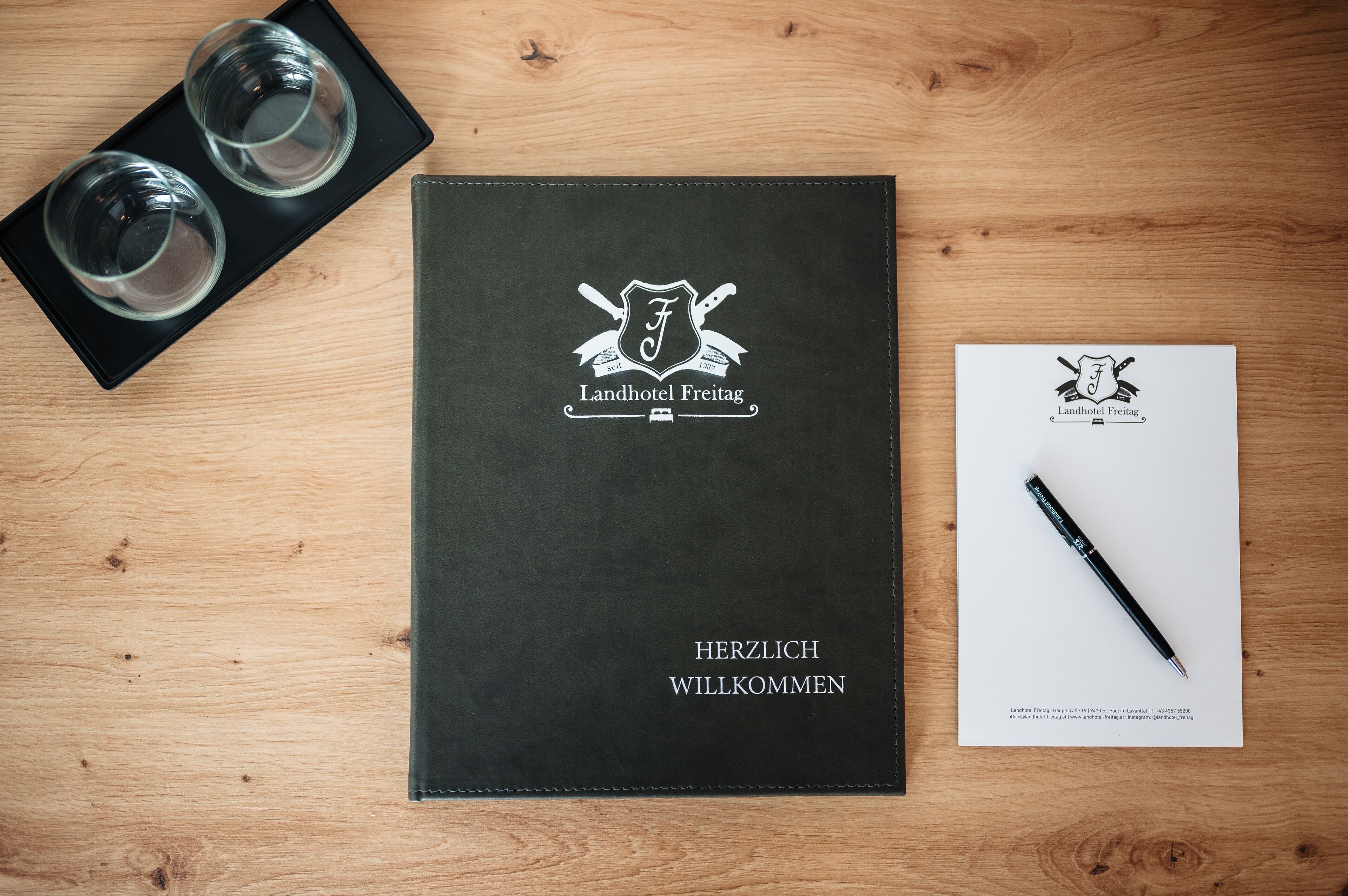Een zwart leren welkomstboekje voor Landhotel Freitag, een pen en twee glazen op een houten tafel.