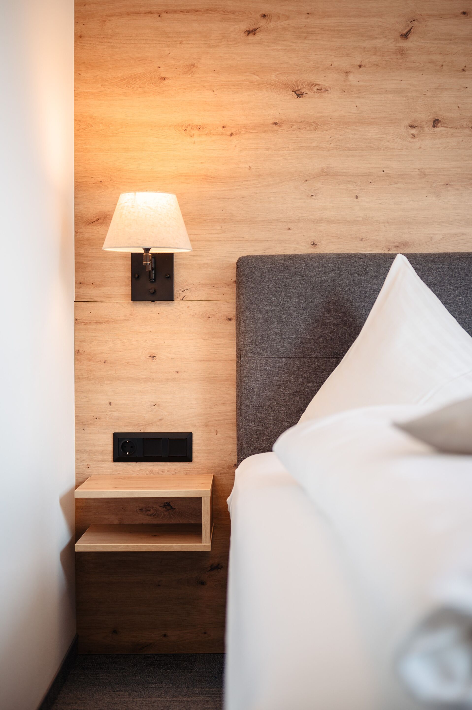 Habitación de hotel ordenada con pared de madera, lámpara de noche, mesita de noche y cama con ropa blanca.