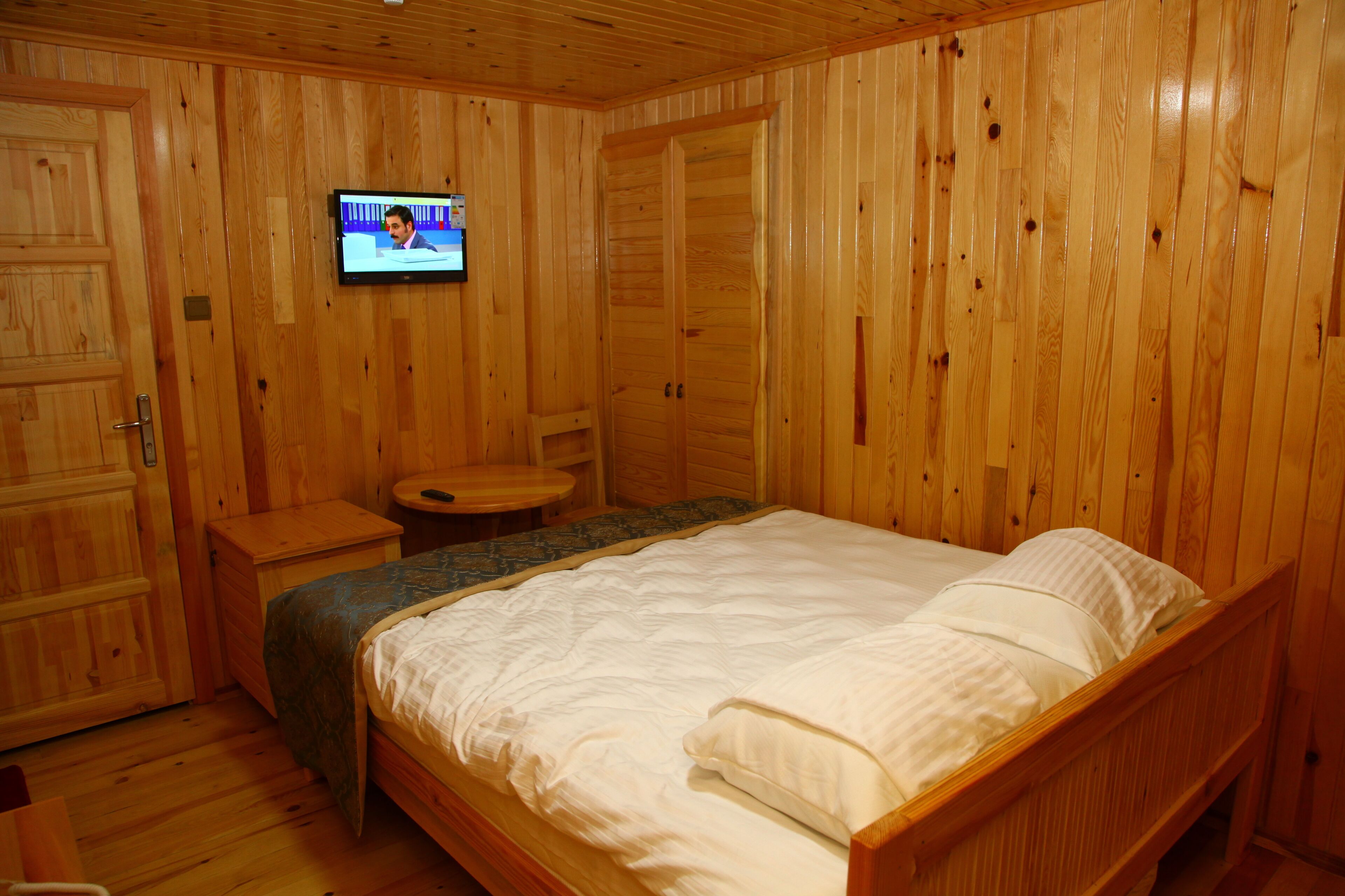 Una habitación con paneles de madera, una cama, un televisor montado en la pared y una mesita redonda.